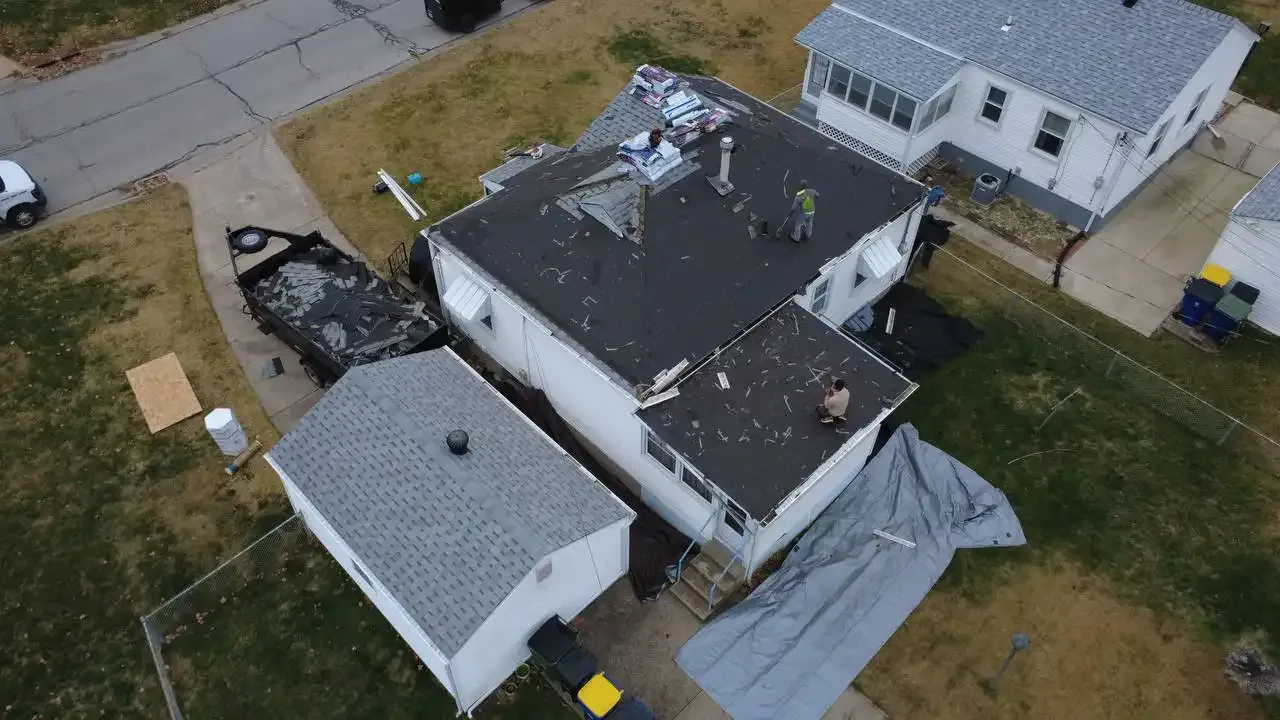 An aerial view of a house with workers on the roof performing repairs. There are materials and debris scattered on the roof and ground, along with trucks parked nearby.