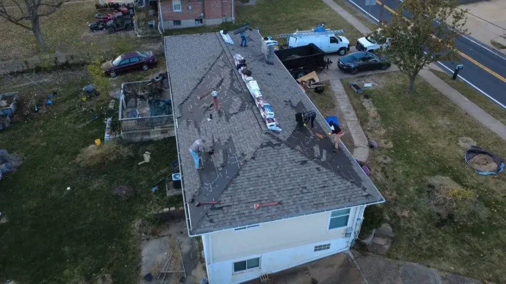 A roofing crew replacing shingles on a large residential home, with a work van, dumpster, and materials staged in the driveway