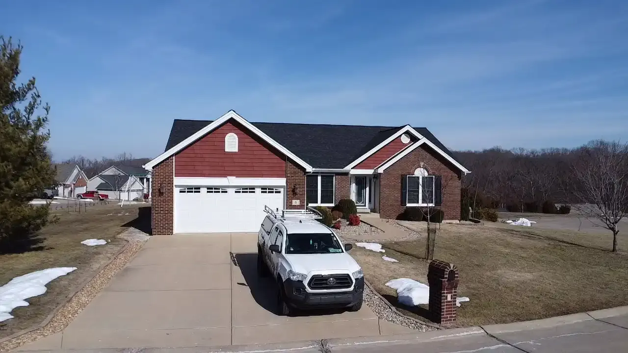 A charming single-story brick house with red siding and a well-maintained lawn, featuring a driveway and a parked white pickup truck in front under a clear blue sky.