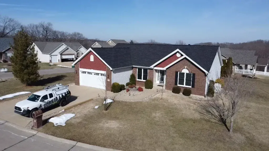 A cozy suburban home with a red and brick exterior, a well-maintained lawn, and a driveway leading to a garage. A service vehicle is parked nearby, and the sky is clear.