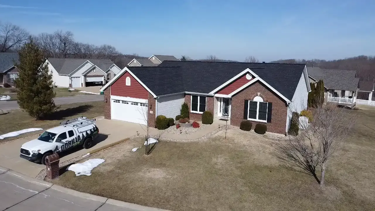A cozy suburban home with a red and brick exterior, a well-maintained lawn, and a driveway leading to a garage. A service vehicle is parked nearby, and the sky is clear.