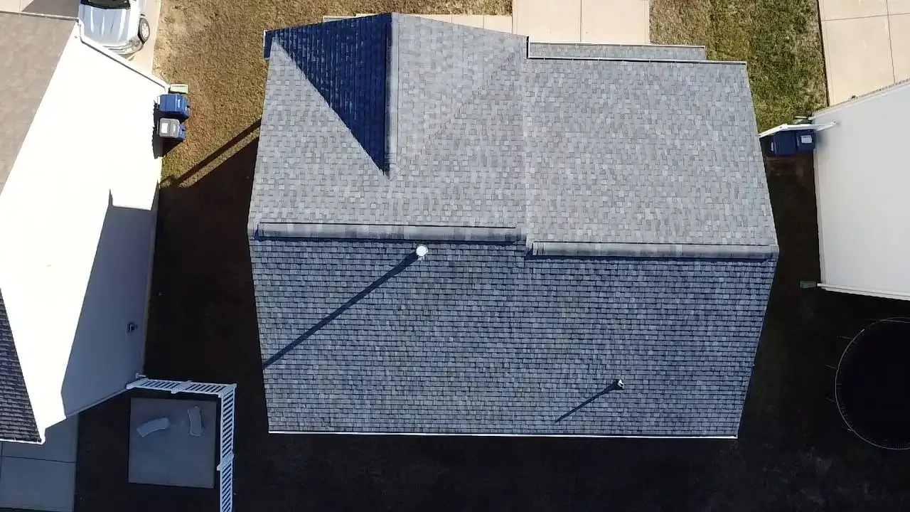Aerial view of a gray shingled roof with a pointed section, showing shadows cast on the surface. The surrounding area includes grassy patches and neighboring rooftops.