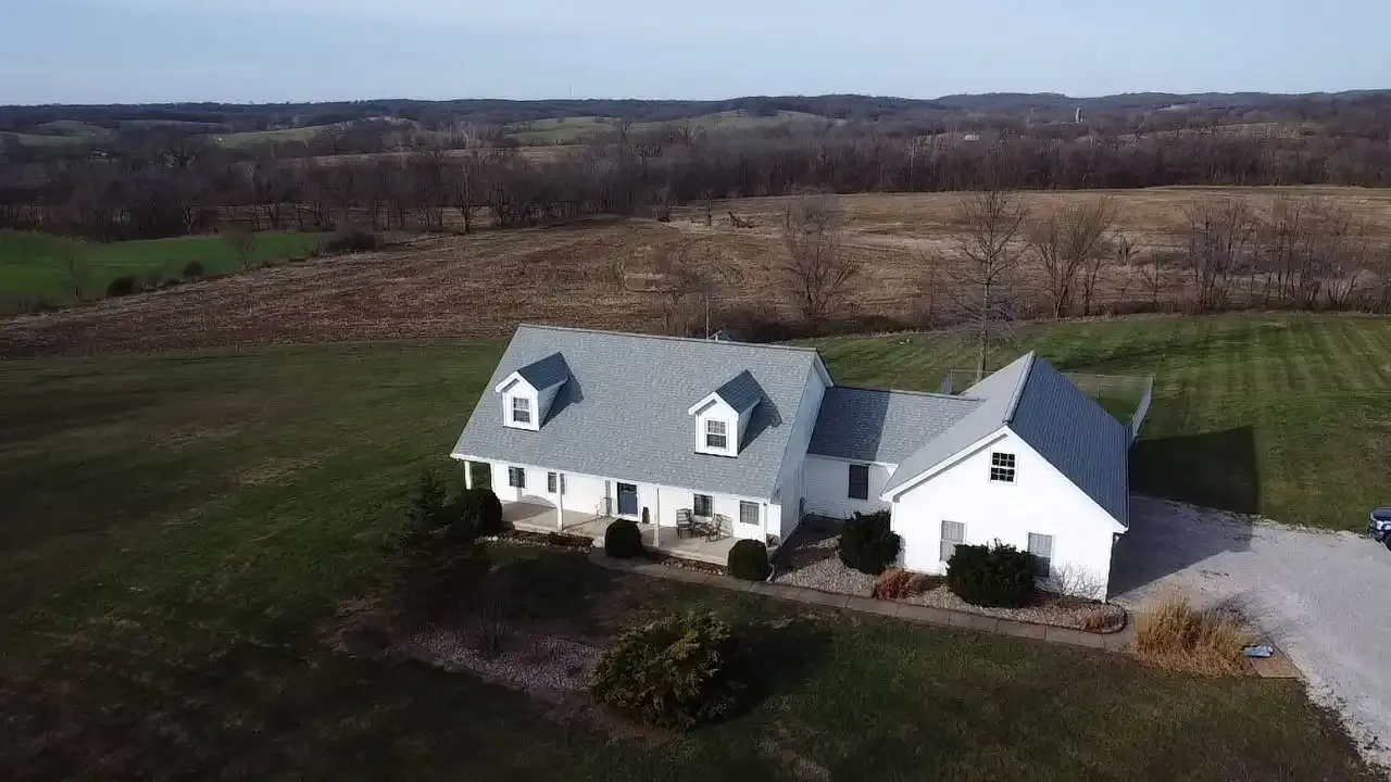 Aerial drone view of white Cape Cod home with gray shingle roof on rural Missouri property with rolling hills.