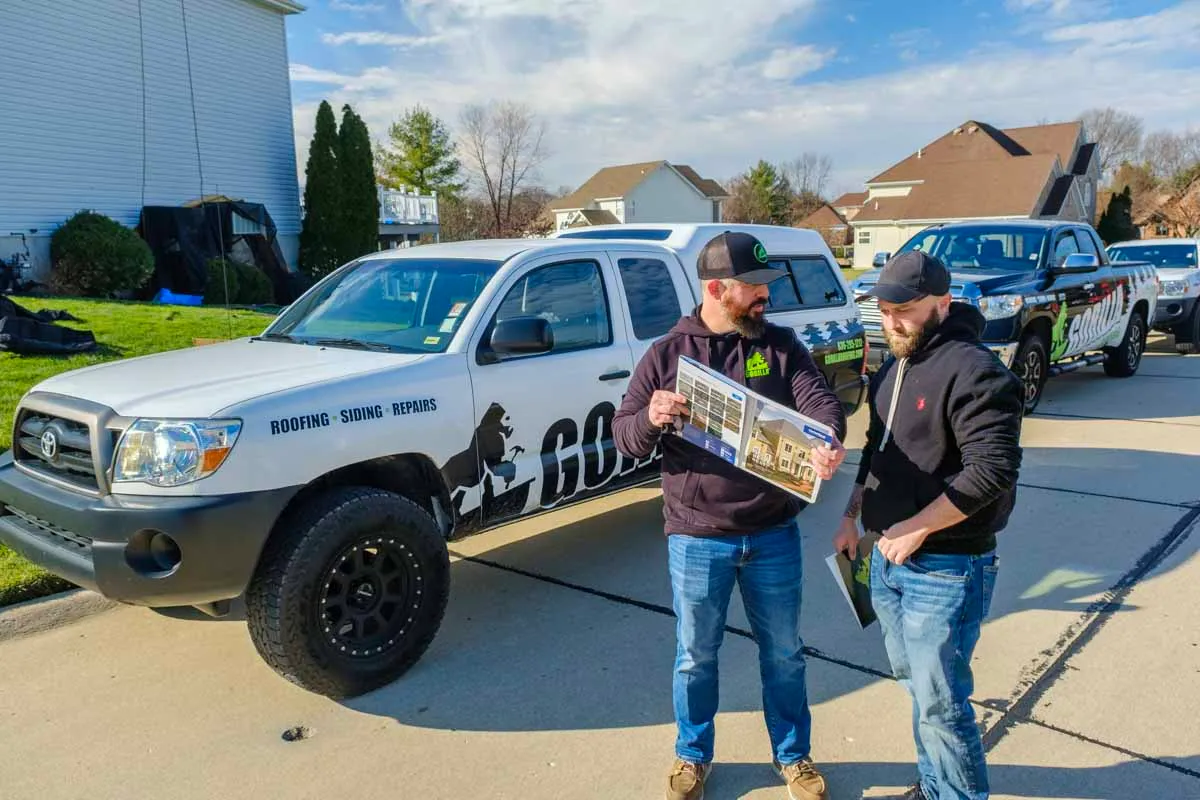 Two Gorilla roofing contractors review project plans beside branded white Toyota Tacoma truck in residential driveway.