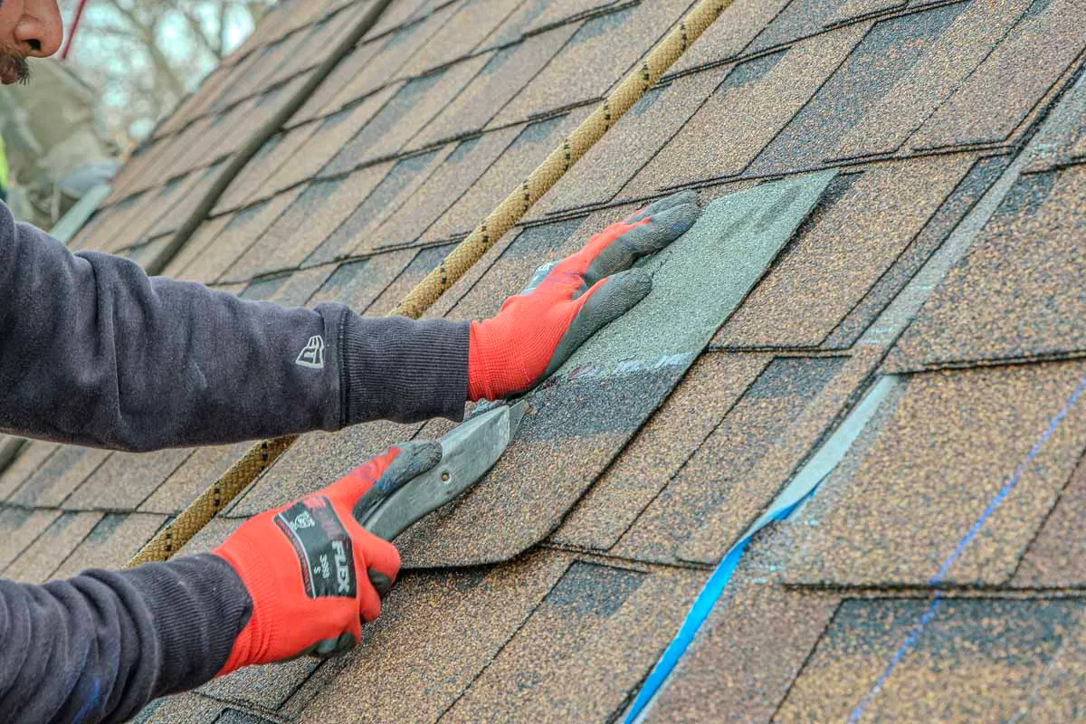 Roofing contractor installing asphalt shingles on a roof, wearing red safety gloves and using a pry tool.
