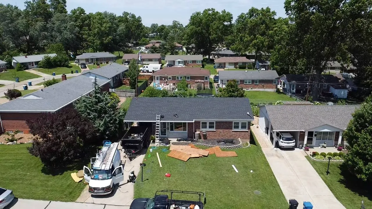 Aerial drone view of a St. Louis residential roof replacement in progress with work van, dumpster, and ladder visible.