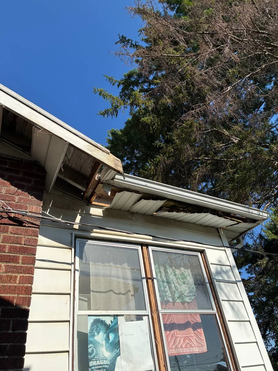 Severely damaged roof soffit and fascia with rotted wood and sagging gutters on a brick home exterior.