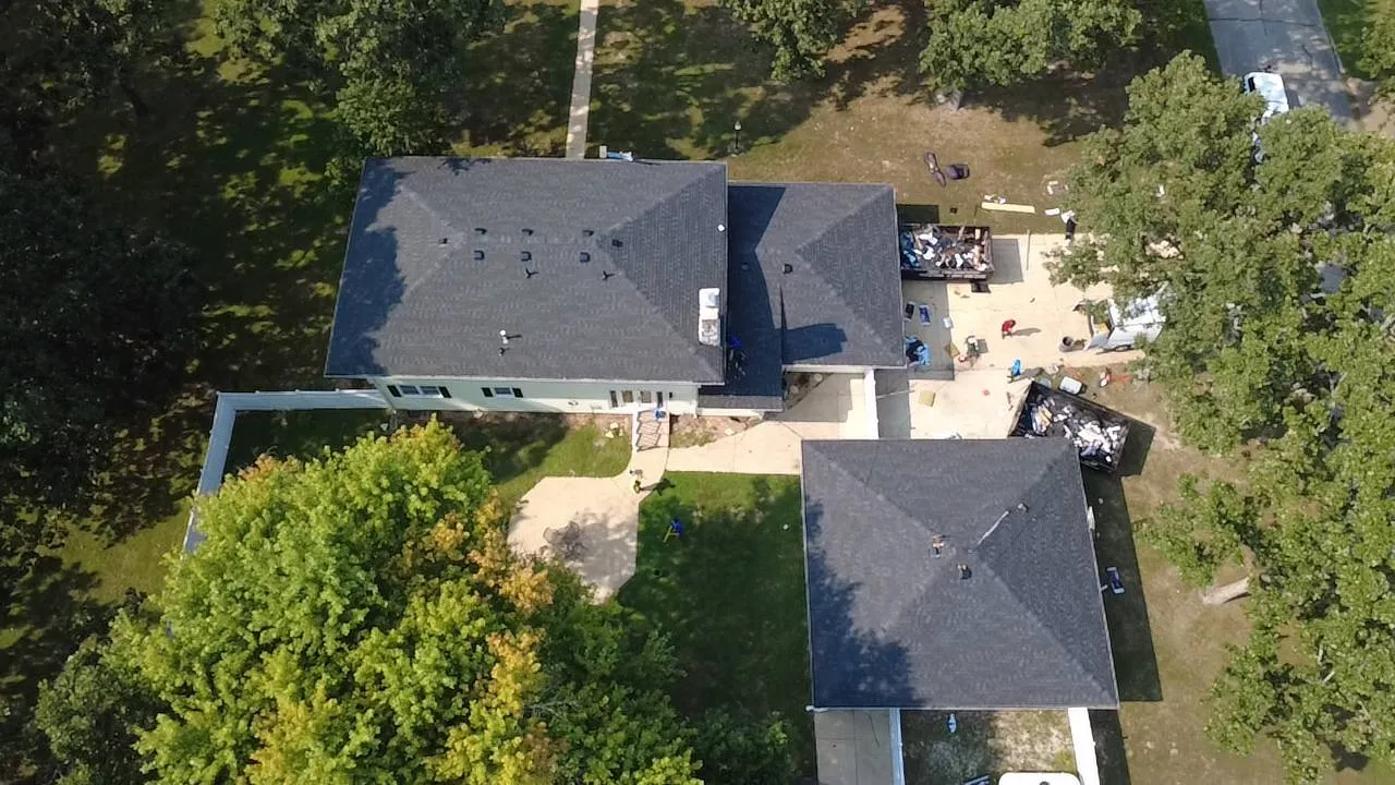 Aerial drone view of residential roof replacement in progress with workers and dumpsters visible on property.