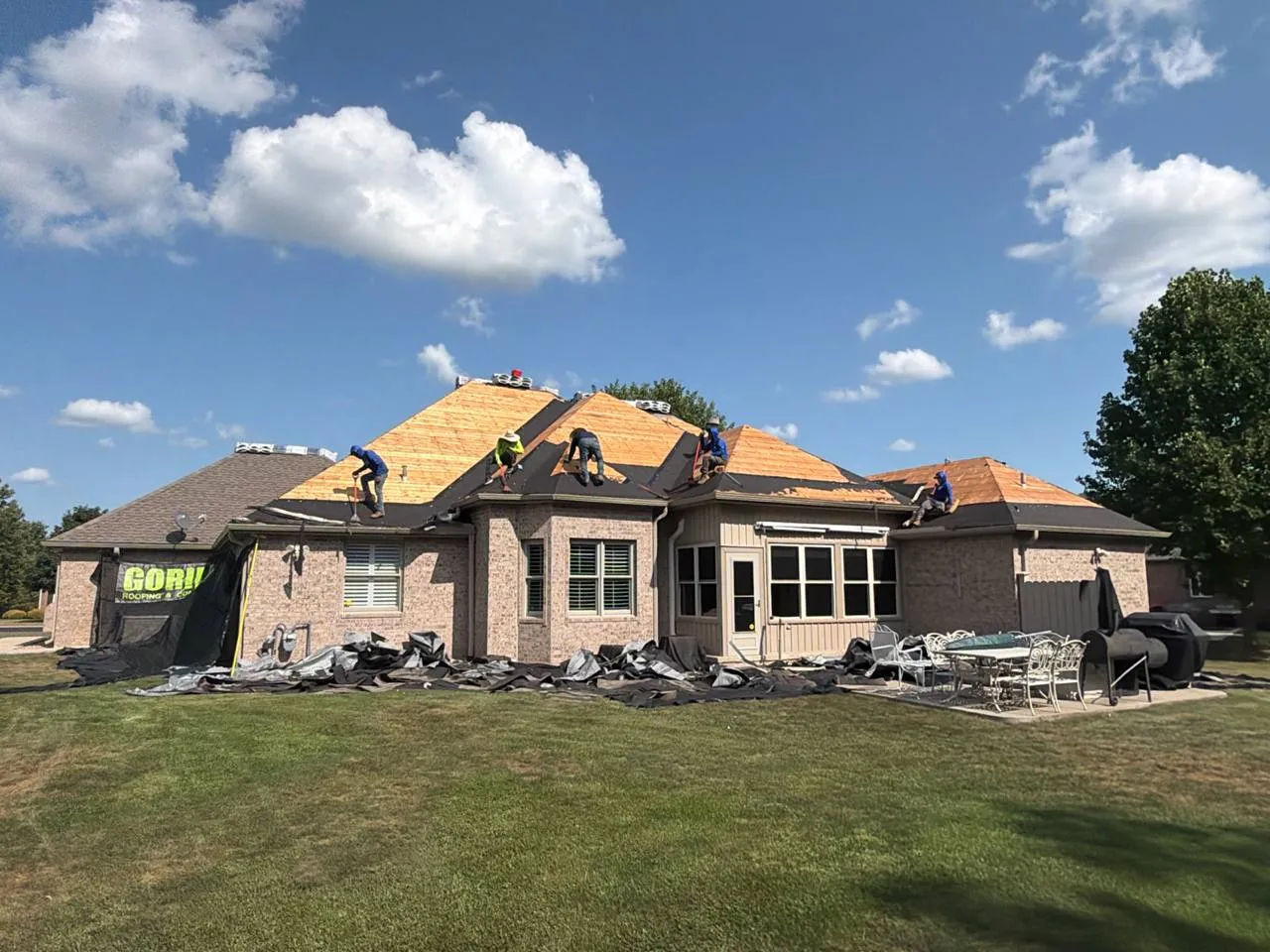 Gorilla Roofing crew replacing shingles on a large brick home, exposing fresh wood decking under blue skies.