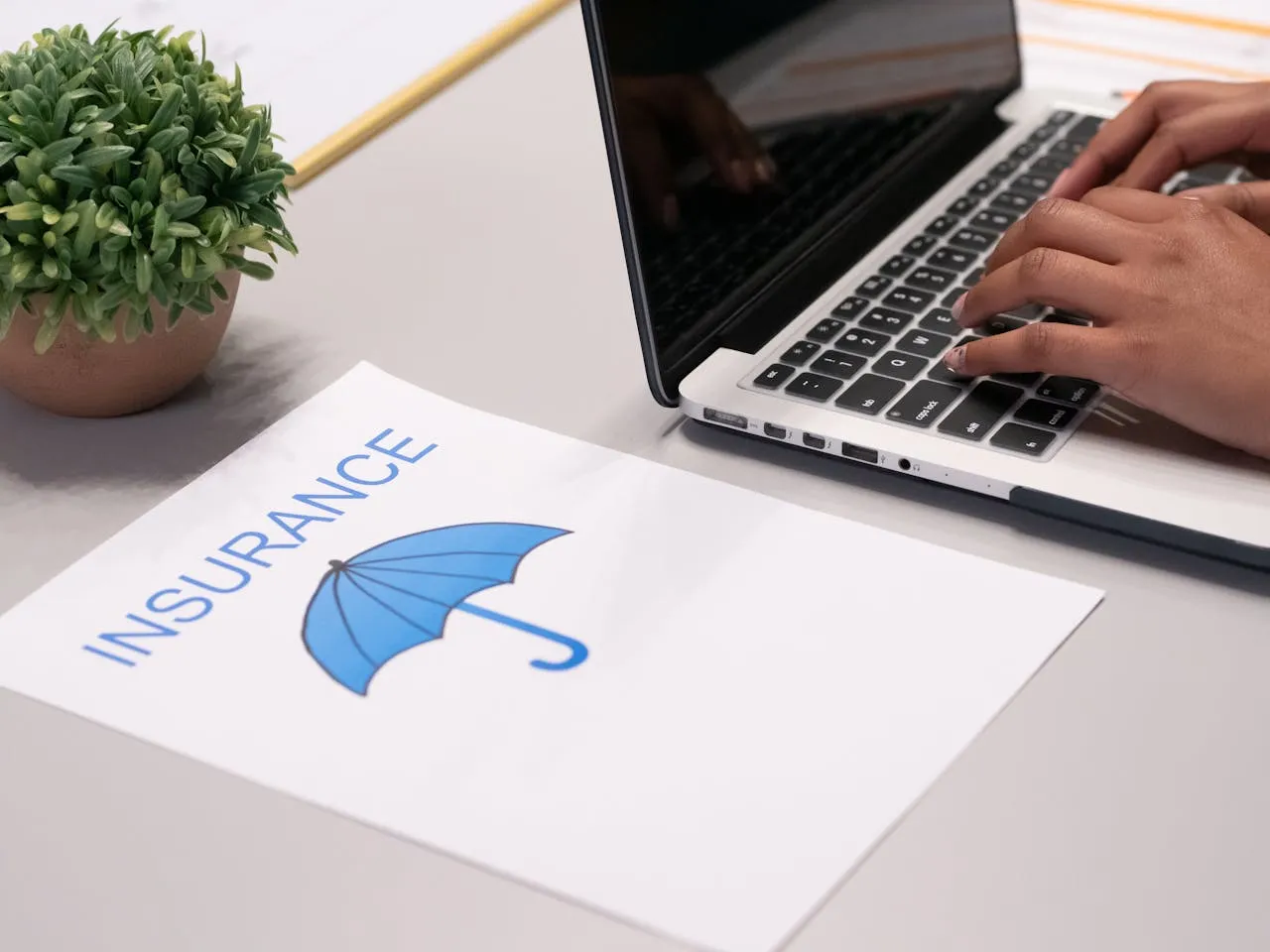 Person typing on laptop next to an insurance document with blue umbrella logo on a desk.