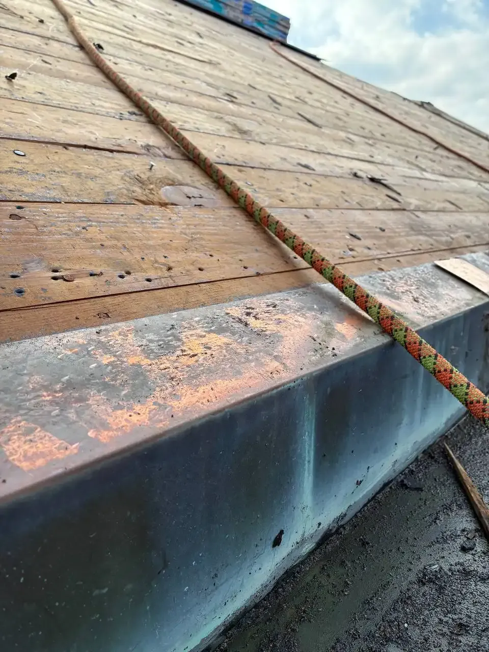 A close-up view of a textured wooden surface on a roof with a colorful climbing rope lying along the edge, set against a cloudy sky.