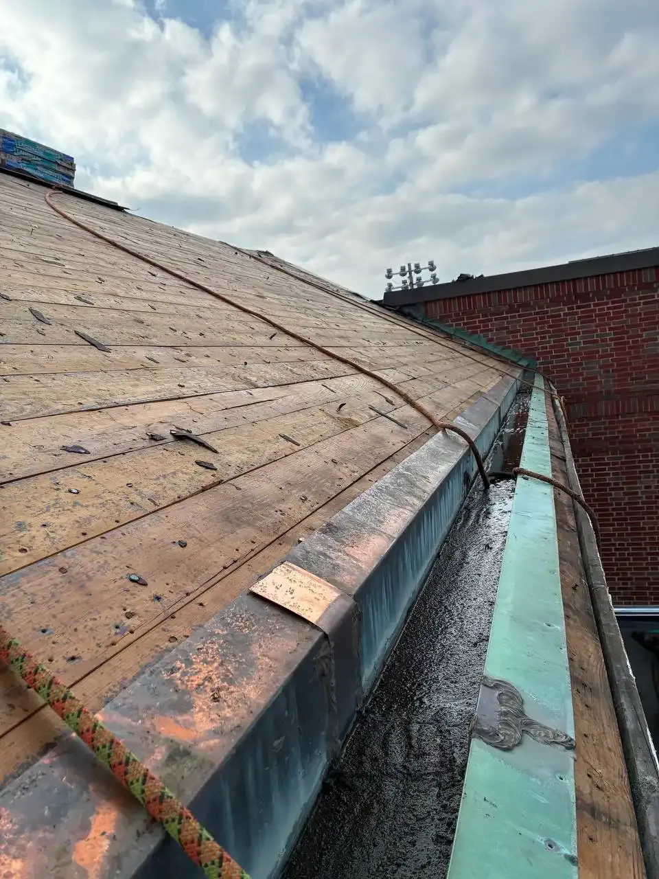 A close-up view of a sloped roof with wooden shingles, showing a gutter along the edge. The sky is partly cloudy in the background.